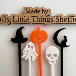 May include: Five Halloween themed acrylic drink stirrers. The stirrers are black, white, and orange. The designs include a witch's hat, a ghost, a pumpkin, a skull, and a crescent moon with a cat sitting on it. The stirrers are made by Crafty Little Things Sheffield.