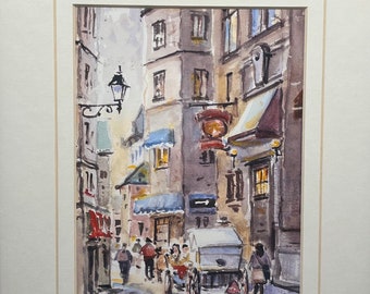 May include: A watercolor painting of a narrow street scene in a European city. The buildings are painted in shades of gray and brown, with some details in blue and red. There are people walking in the street, and a horse-drawn carriage is parked in the middle of the street. The painting is signed "J.R.Lobbe" in the lower right corner.
