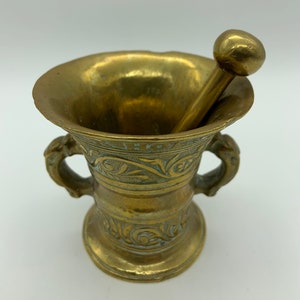 May include: A brass mortar and pestle with ornate floral designs. The mortar has two handles and a round base. The pestle has a round head.