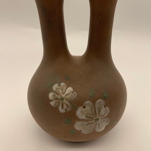 May include: A brown ceramic vase with two spouts and a handle. The vase is decorated with white flowers and green leaves.