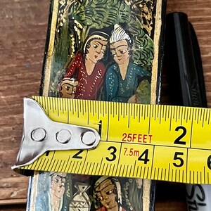 May include: A black wooden box with intricate gold and colorful painted designs. The box features a scene with two figures in traditional clothing, surrounded by floral motifs. The box is being measured with a tape measure, showing a length of 3.75 inches.