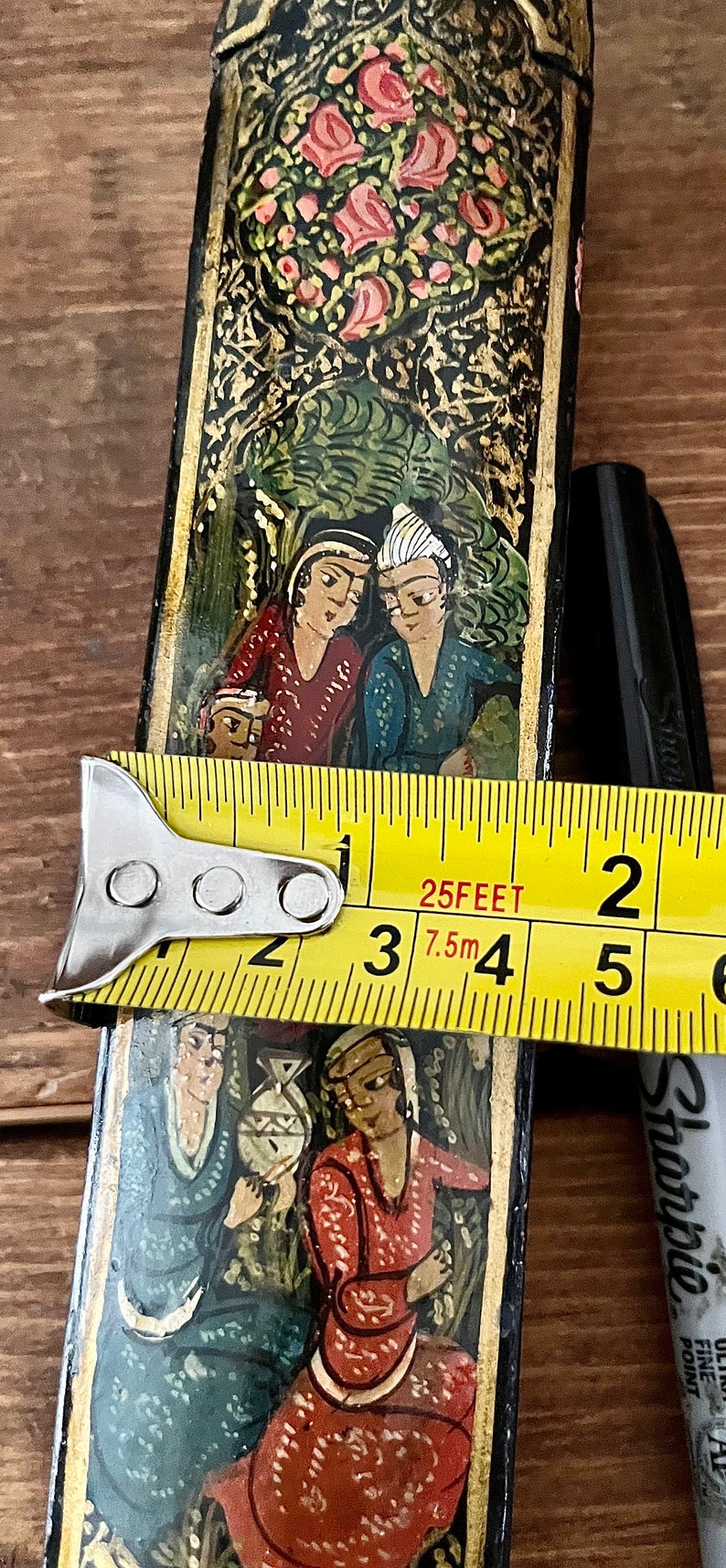 May include: A black wooden box with intricate gold and colorful painted designs. The box features a scene with two figures in traditional clothing, surrounded by floral motifs. The box is being measured with a tape measure, showing a length of 3.75 inches.