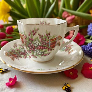 May include: A white teacup and saucer set with a floral design featuring red berries, green leaves, and a red and gold ribbon pattern. The cup and saucer have gold trim. The set is on a white surface with flowers.