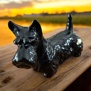 May include: Black ceramic figurine of a Scottish Terrier dog. The figurine is lying down with its head turned to the side. The dog has a shiny black finish and is detailed with fur.