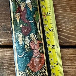May include: A black and gold rectangular box with a painted scene of people in traditional clothing. The box is decorated with floral patterns and has a hinged lid. The box is 8 inches long.