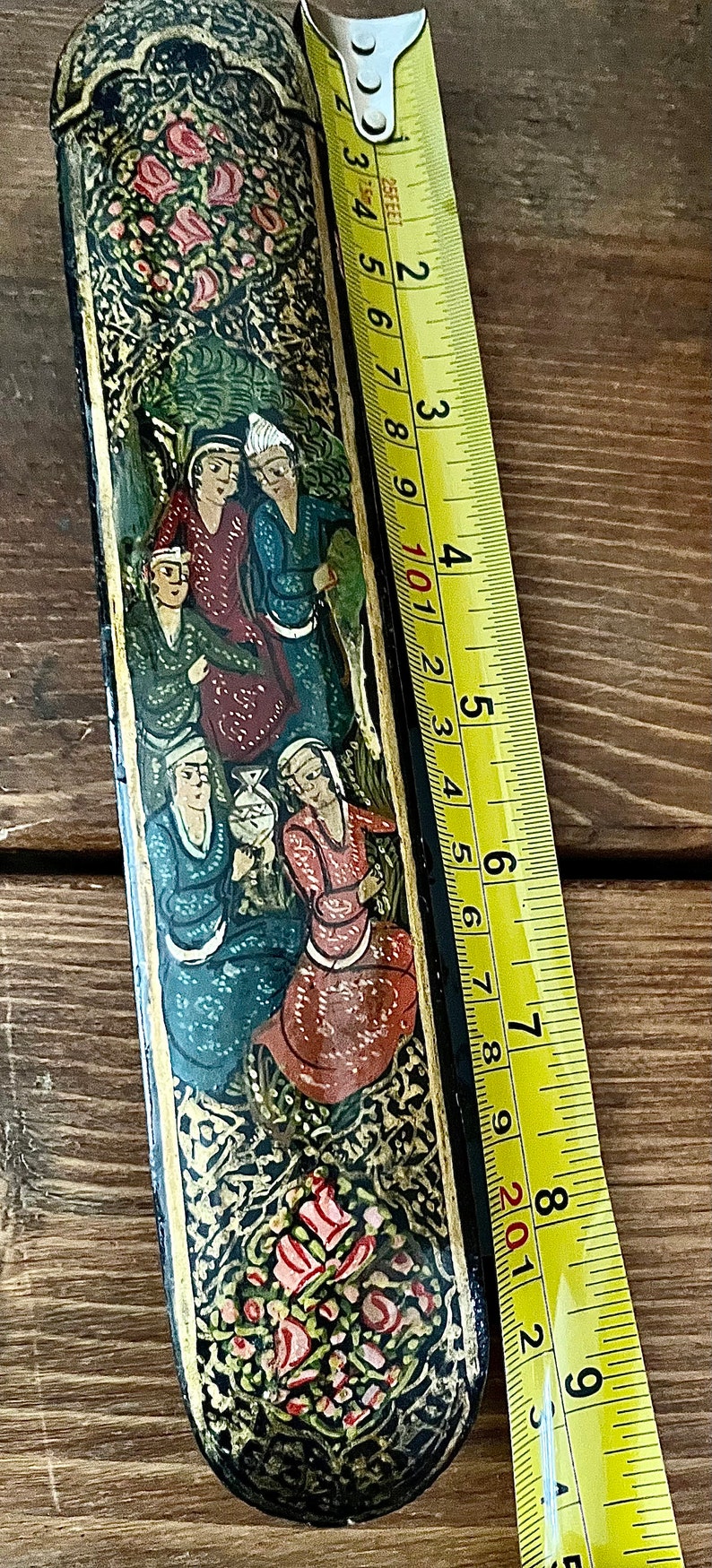 May include: A black and gold rectangular box with a painted scene of people in traditional clothing. The box is decorated with floral patterns and has a hinged lid. The box is 8 inches long.