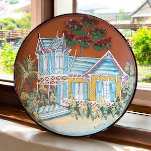 May include: A hand-painted ceramic plate featuring a colourful house with a blue roof, yellow walls, and blue shutters. The house is surrounded by palm trees and lush greenery. The plate is signed "Robbider".