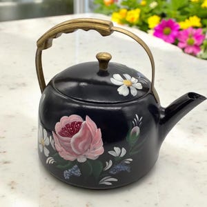May include: A black teapot with a brass handle and painted floral designs. The teapot features a pink rose, white daisies, and green leaves.