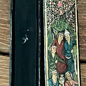 May include: A black wooden box with intricate gold and floral designs. The box is open, revealing a black interior. The lid features a painted scene of people in a garden setting.