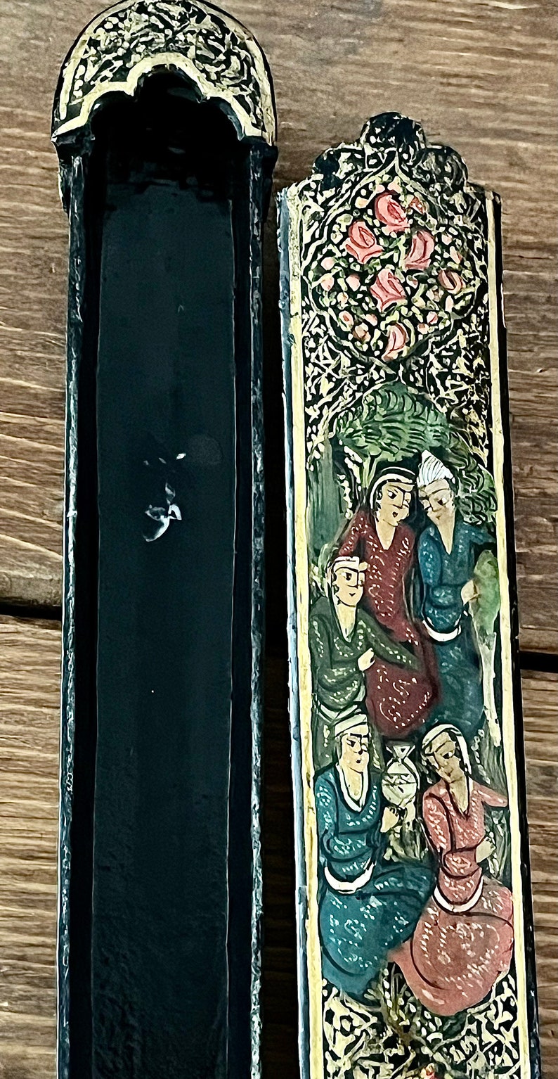May include: A black wooden box with intricate gold and floral designs. The box is open, revealing a black interior. The lid features a painted scene of people in a garden setting.