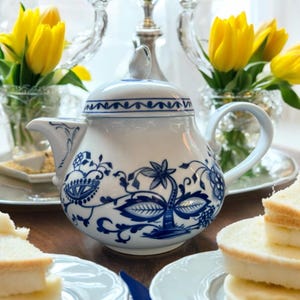 Op de afbeelding: Een witte theepot met een blauw en wit bloemenpatroon. De theepot heeft een deksel en een handvat. De theepot staat op een houten tafel.