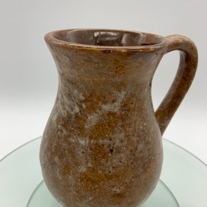 May include: A handmade brown ceramic pitcher with a handle. The pitcher has a bulbous body and a wide mouth. The surface has a speckled, textured appearance with lighter brown and white accents. The pitcher is likely used for serving beverages.