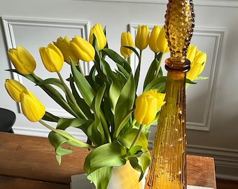 May include: A tall amber glass bottle with a textured design and a pointed top. The bottle is filled with a clear liquid. A bouquet of yellow tulips is in a white vase in the foreground.
