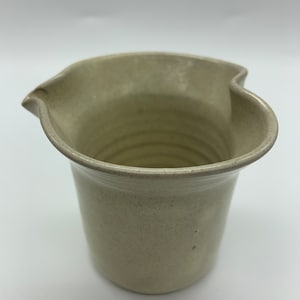 May include: A light beige ceramic jug with a unique heart-shaped lip. The jug has a smooth, slightly textured surface and a slightly flared base.