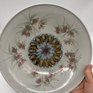 May include: A white ceramic bowl with a floral design. The bowl has a center design of brown and blue petals with a pink flower in the center. The rim of the bowl is decorated with pink flowers and green leaves.