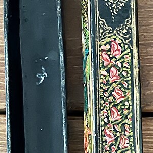 May include: A black wooden box with intricate gold and floral designs. The box is open, revealing a black interior. The box is approximately 10 inches long.