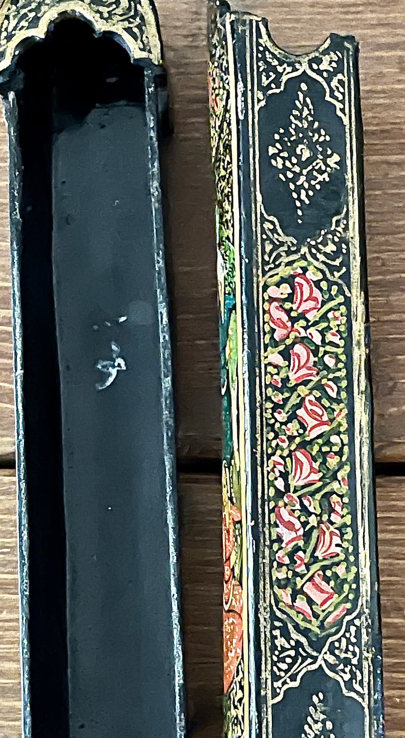 May include: A black wooden box with intricate gold and floral designs. The box is open, revealing a black interior. The box is approximately 10 inches long.