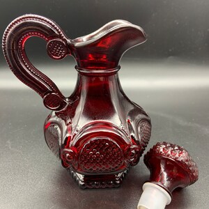 May include: A red glass jug with a decorative handle and a matching stopper. The jug has a textured, diamond-shaped pattern.