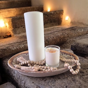 May include: A white pillar candle and a smaller candle with a brown wax top are displayed on a brown ceramic plate with a beaded garland. The candles are on a set of stone steps.