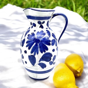 May include: A white ceramic jug with a blue floral design and a blue rim. The jug has a handle and is sitting on a white surface.