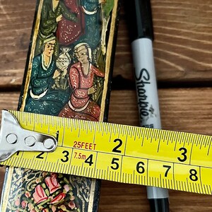 May include: A black and gold rectangular box with a floral design and painted figures. The box is approximately 4 inches long.