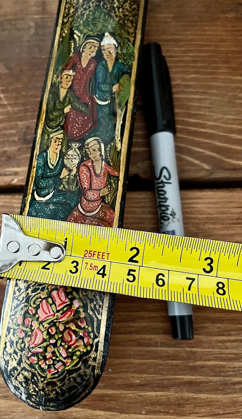 May include: A black and gold rectangular box with a floral design and painted figures. The box is approximately 4 inches long.