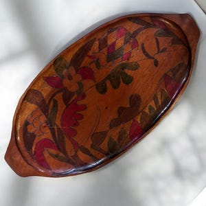May include: A wooden serving tray with a hand-painted floral design in shades of red, green, and brown. The tray has two handles and is oval shaped.