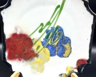 May include: A white ceramic plate with a black rim and a floral design in shades of red, yellow, blue, and green. The plate is on a wooden stand.