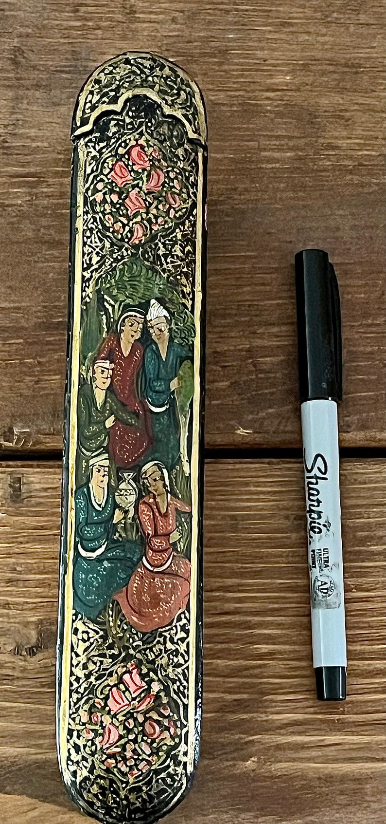 May include: A black and gold rectangular box with a floral design and a scene of people in traditional clothing. The box is decorated with intricate details and has a hinged lid.