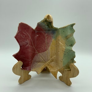 Peut inclure: Un plat en céramique en forme de feuille avec un glaçage rouge, vert et jaune. Le plat est sur un support en bois.