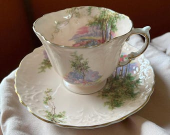 May include: A vintage teacup and saucer set. The white porcelain cup and saucer feature a landscape scene with trees and a pink and blue sky. The edges are scalloped and trimmed with gold.