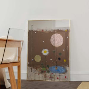Puede incluir: Impresión artística abstracta con un fondo marrón, un gran círculo rosa punteado y una margarita blanca. También se ven un pequeño estanque azul y un círculo amarillo. La obra de arte está enmarcada en dorado y se apoya en una pared blanca.