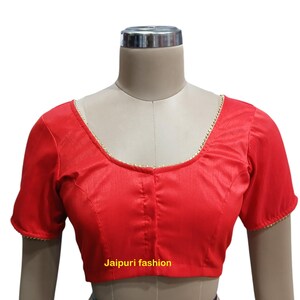 May include: Red sleeveless blouse with a gold trim around the neckline and sleeves. The blouse is on a mannequin. The text "Jaipuri fashion" is printed below the blouse.
