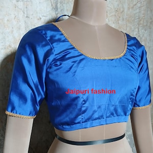 Può includere: Una blusa in satin blu reale con bordini dorati intorno al collo e alle maniche. La blusa ha una scollatura rotonda e maniche corte. Il testo "Jaipuri fashion" è stampato sul davanti della blusa.