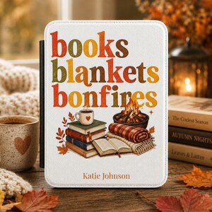 May include: A white e-reader case featuring the text books blankets bonfires in orange, red, and brown. The design includes a stack of books, a steaming mug with a heart, a small bonfire, a plaid blanket, and the name Katie Johnson at the bottom.