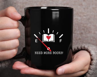 May include: Black ceramic mug with a white graphic of a fuel gauge, a book with a heart, and the text "NEED MORE BOOKS!". The gauge needle points towards empty. The mug is held by a person.