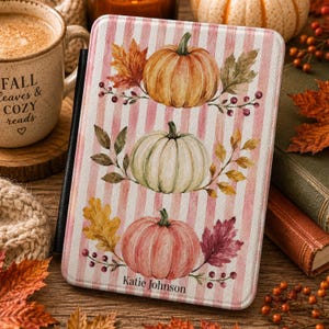 May include: A rectangular e-reader case featuring a light pink and white vertical striped background. The design displays three watercolor-style pumpkins in orange, cream, and muted pink, surrounded by autumn leaves and berries. The name Katie Johnson is printed at the bottom.