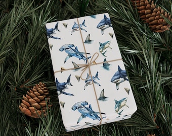 May include: A wrapped gift with shark-themed wrapping paper. The paper features various blue shark illustrations on a white background, tied with twine. The gift is surrounded by pine needles and pine cones.