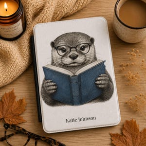 May include: A white e-reader cover featuring a detailed black and white illustration of an otter wearing glasses and reading a blue book. The name Katie Johnson is printed in black text at the bottom of the cover, which has a black spine.
