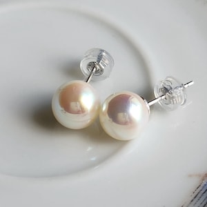 May include: A pair of pearl stud earrings. The earrings feature lustrous, round, off-white pearls with a subtle iridescent sheen. The posts and backs are silver-toned. The earrings are displayed on a white surface.