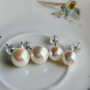 May include: Four white pearl stud earrings with iridescent sheen, displayed on a white plate. The earrings vary in size, with clear plastic backings. The pearls have a soft, lustrous appearance, reflecting light.