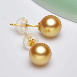 May include: A pair of gold-colored pearl stud earrings with clear, plastic earring backs. The earrings have a post and a round, lustrous pearl. The earrings are displayed on a white surface.