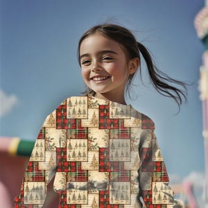 May include: A young person smiles, wearing a long-sleeved shirt. The shirt features a patchwork design with Christmas tree and plaid patterns in red, green, and beige. The background is a clear blue sky.