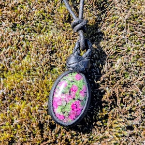 Könnte beinhalten: Ovale Anhänger Halskette mit einem polierten Stein mit rosa und grünen Farbtönen, eingefasst in einen schwarzen Lederrahmen. Der Anhänger hängt an einer schwarzen Lederschnur, die oben verknotet ist.