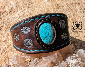 May include: A brown leather cuff bracelet with turquoise stitching and a large oval turquoise stone. The bracelet features embossed paw prints and other designs. The brand logo "Valea Gems" is visible in the background.