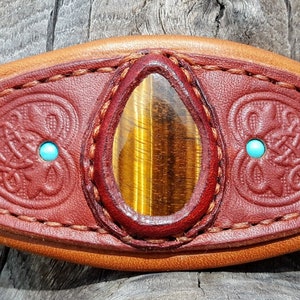 Op de afbeelding: Een bruin lederen halster voor paarden met een tijgeroog-steen en turquoise accenten. Het halster heeft een Keltisch knoopontwerp.
