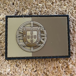 Peut inclure: Un patch en tissu brun et beige avec les armoiries portugaises au centre. Le patch est fixé à un mur en pierre.