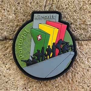 May include: Airsoft patch with a green, yellow, and red design. The patch features a silhouette of soldiers and the text "Airsoft Portugal".