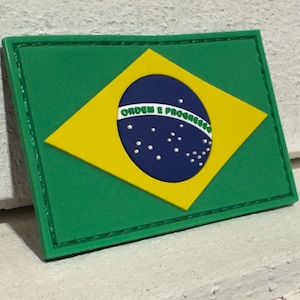 Puede incluir: Un parche rectangular verde con la bandera brasileña. La bandera tiene un rombo amarillo, un círculo azul con estrellas blancas y el texto "ORDEM E PROGRESSO".