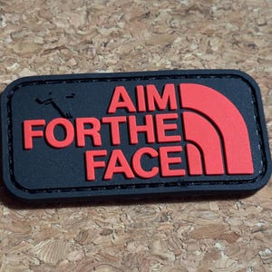 Op de afbeelding: Een zwart rechthoekig embleem met de rode woorden "AIM FOR THE FACE". De patch heeft een zwarte rand en een rood logo dat lijkt op een bergketen. De achtergrond is een getextureerd, lichtbruin oppervlak.